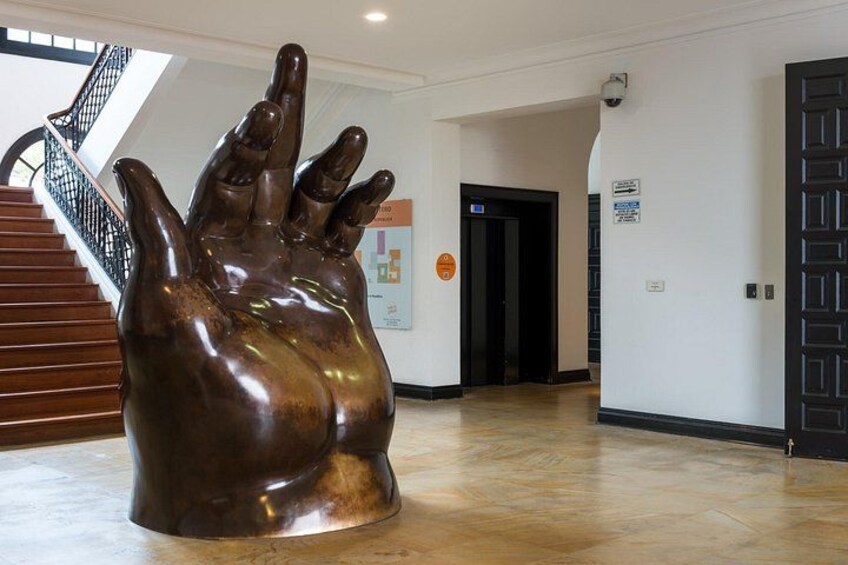 A large bronze sculpture of a hand in a hallway with wooden floors, a staircase, and a door in the background. 4