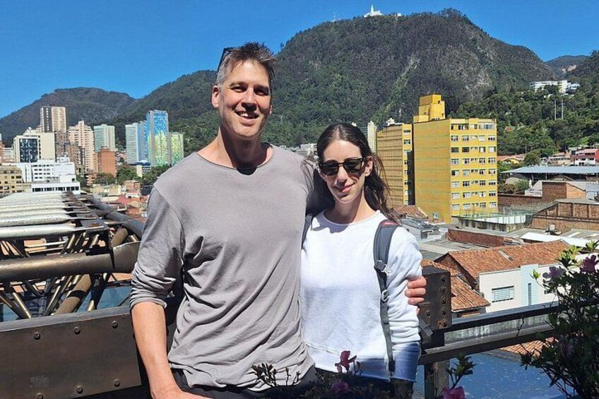A man and woman standing on a bridge with a cityscape and mountains in the background. 1