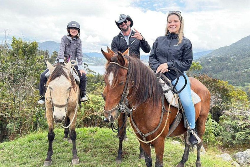 Three people riding horses on a grassy hill with a mountainous landscape in the background. 1