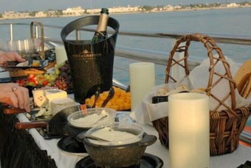A table with food, a bottle of champagne, and a candle by the water. 1