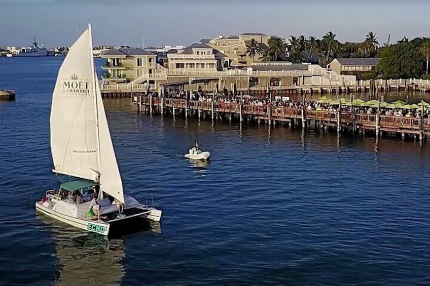 A sailboat with a white sail and green lettering on the water, docked at a pier with a crowd of people and buildings in the background. 2