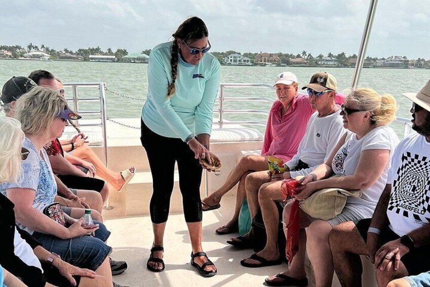 A group of people on a boat, some seated, some standing, with a person in a light blue shirt leading a discussion. 3