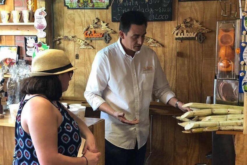 A person in a white chef's coat standing at a counter with a woman in a patterned dress. There are wooden shelves with various items and a chalkboard with writing in the background. 4