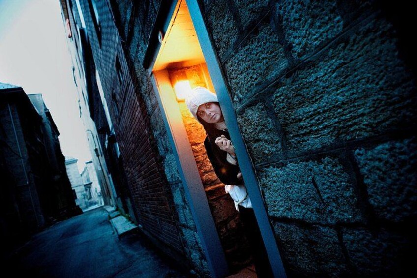 Une ruelle étroite aux murs de pierre, une personne qui jette un coup d’œil depuis une porte, et un lampadaire qui éclaire la scène. 1