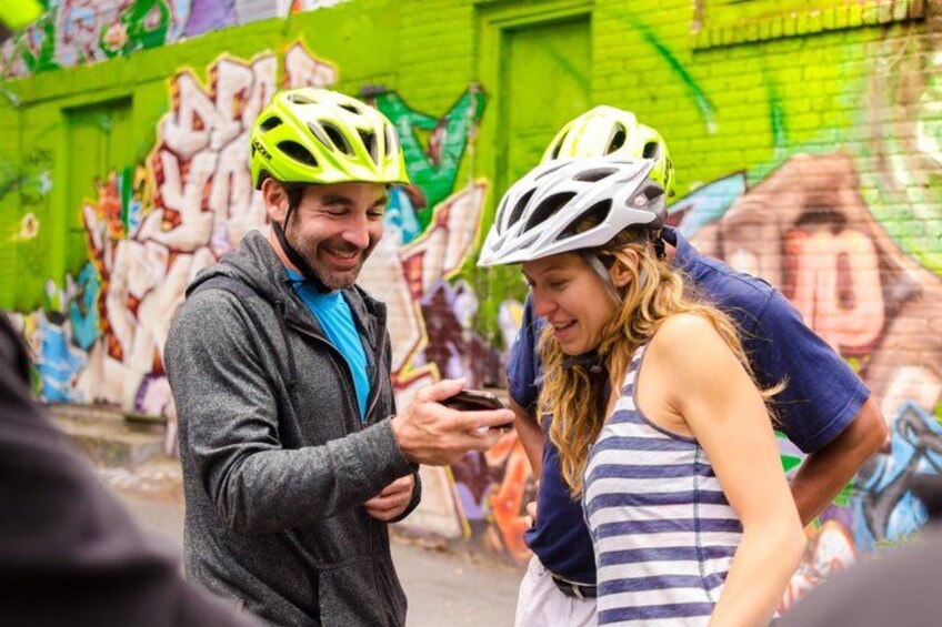 Two people wearing helmets, one in a grey jacket and the other in a striped tank top, standing in front of a graffiti-covered wall. 2