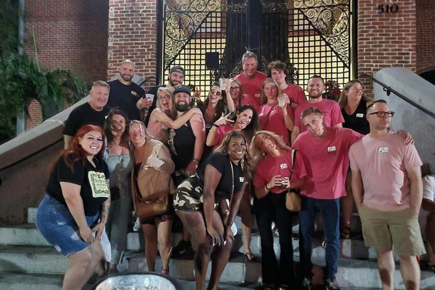 A group of people in red shirts gathered on steps with a brick building and decorative metal gates in the background. 2
