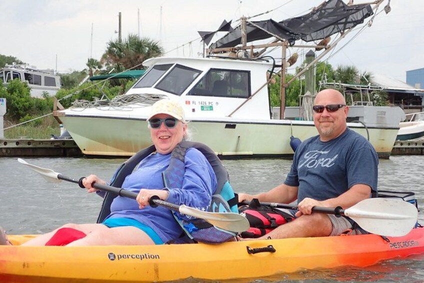 Two people kayaking on a river with boats docked in the background. 3