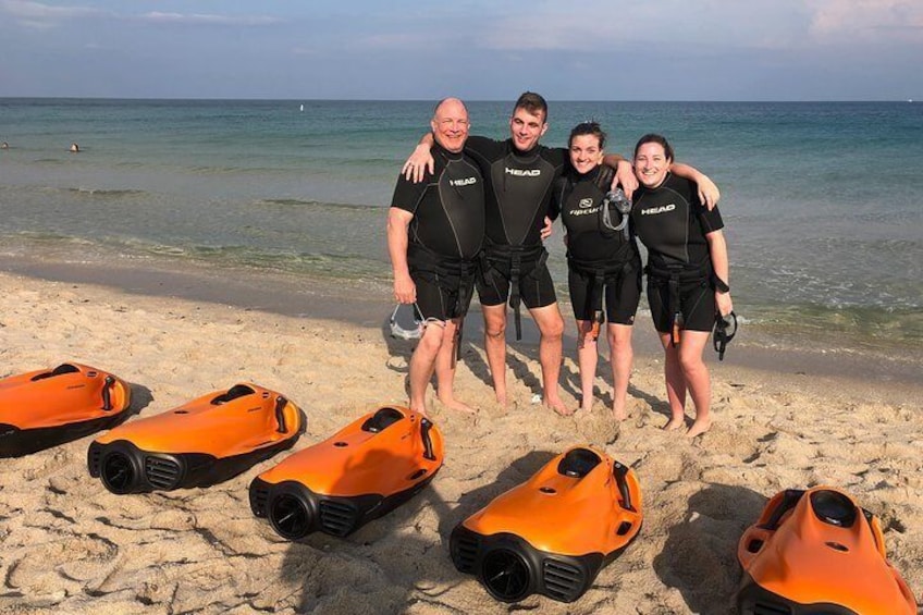 Fire personer i våddragter står på en strand med orange jetski i forgrunden. 4