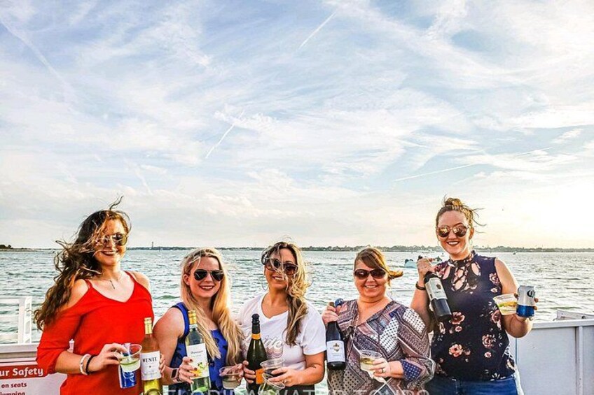 A group of people on a boat with bottles and glasses, a body of water, and a distant shoreline. 3