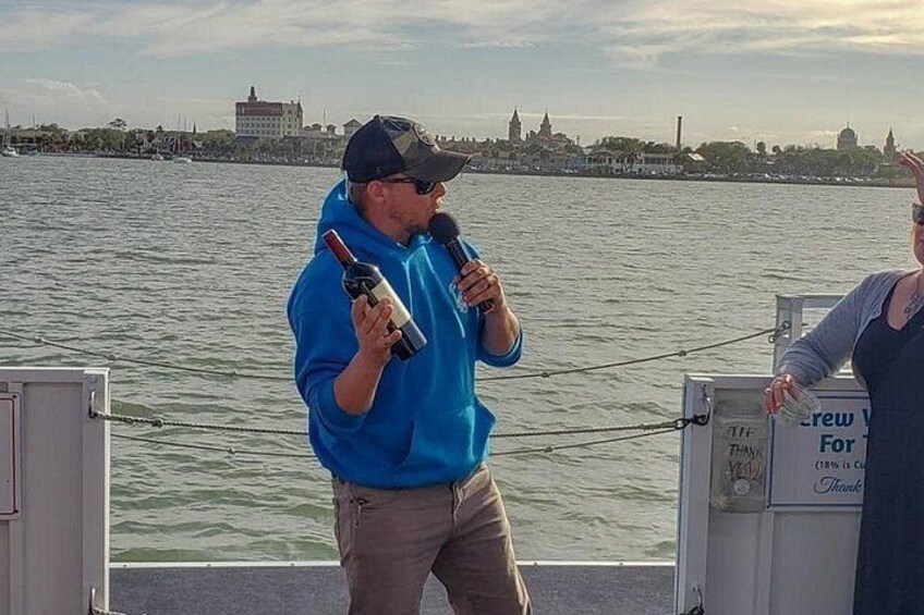 A person holding a microphone and a bottle, standing by a waterfront with a cityscape in the background. 5