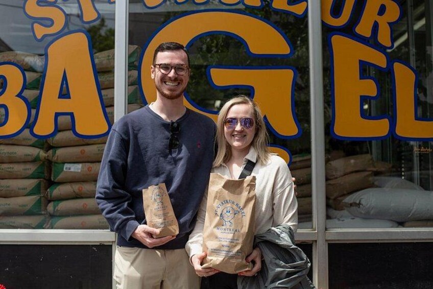 Two people standing in front of a store window with large yellow letters spelling 'ST. BARTOUL BAGEL'. 2
