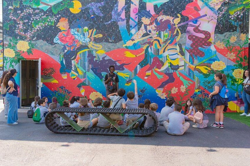 Ein Wandgemälde mit leuchtenden Farben, um das sich eine Gruppe von Kindern und Erwachsenen versammelt hat. 3