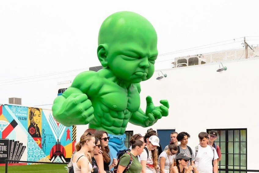 Eine große, grüne Statue einer kräftigen Gestalt, eine Menschenmenge und ein farbenfrohes Wandbild im Hintergrund. 5