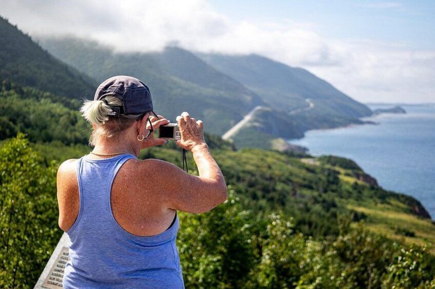 A person taking a photo of a coastal landscape with mountains and greenery. 2