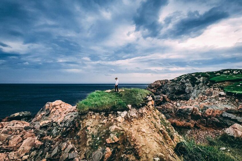 A rocky coastline with a person standing on a grassy cliff overlooking the sea. 2