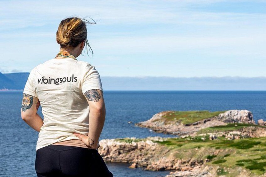 A person standing by the sea with a tattooed arm, wearing a white t-shirt with 'vibingsouls' on the back, and a rocky coastline in the background. 2