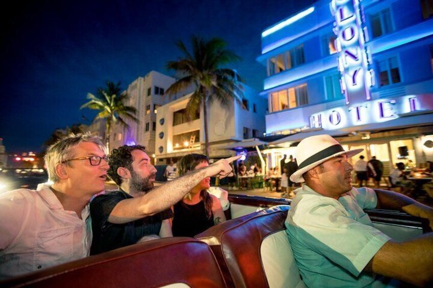A group of people in a convertible car at night, with a hotel in the background. 1