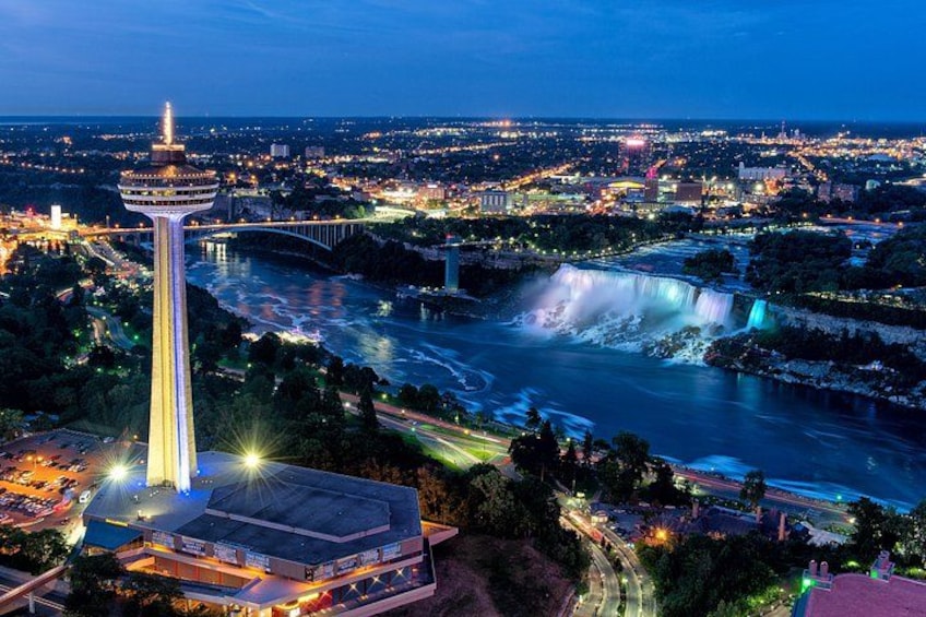 A cityscape at night with a prominent tower, a waterfall, and a bridge. 1