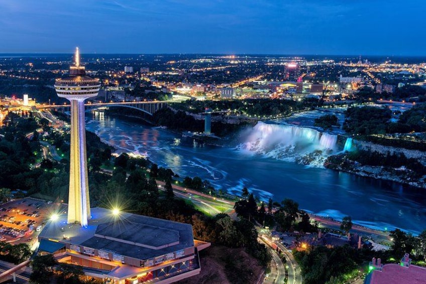 A cityscape at night with a prominent tower, a waterfall, and a bridge. 1