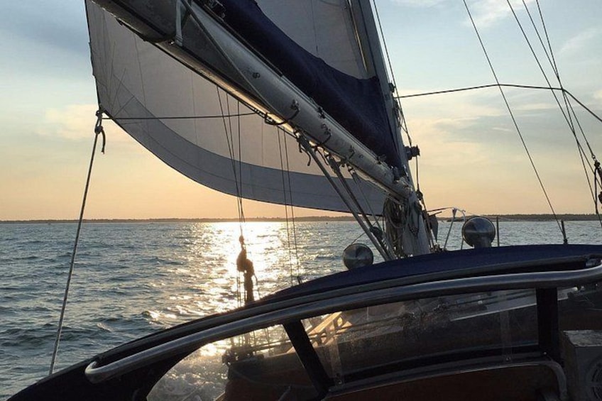 A sailboat with a person standing on the deck, a sunset in the background, and calm water. 2