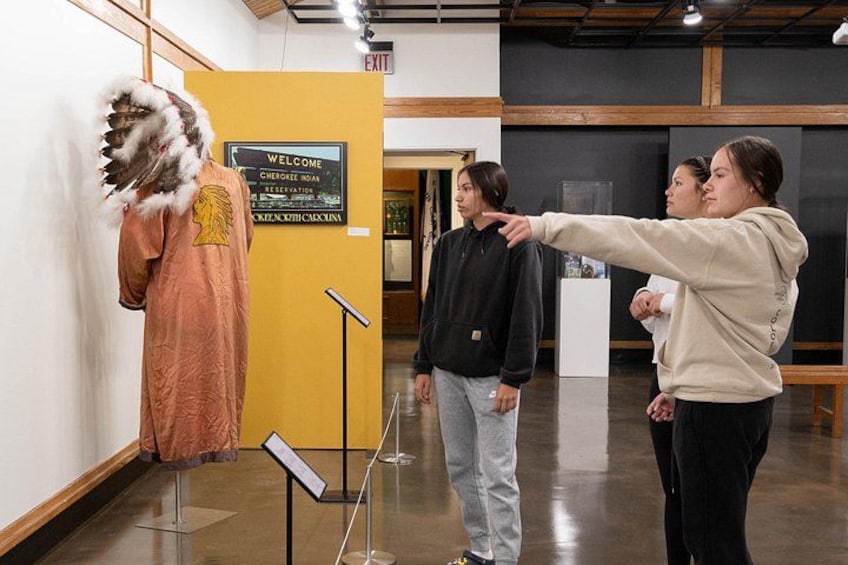 Een binnengalerij met kunst, een etalagepop gekleed in traditionele kleding, een bord met de tekst 'Welcome Cherokee Nation Reservation' en een groep mensen die de tentoonstelling bekijken. 4
