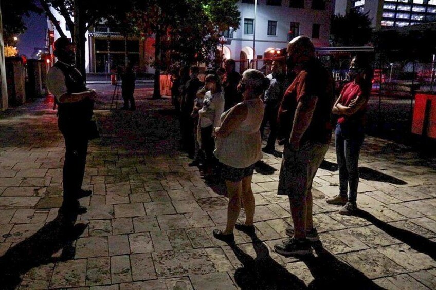 Escena nocturna al aire libre con gente de pie en un área pavimentada, posiblemente una plaza pública o calle. 4