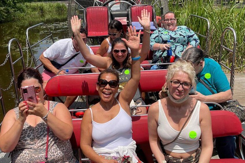 People on a boat with red seats, some raising their hands, and a calm water body in the background. 3