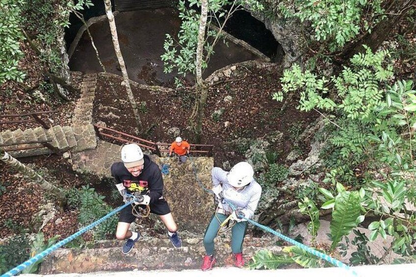 Two people rappelling down a rocky cliff with a rope, surrounded by greenery and a cave entrance. 2