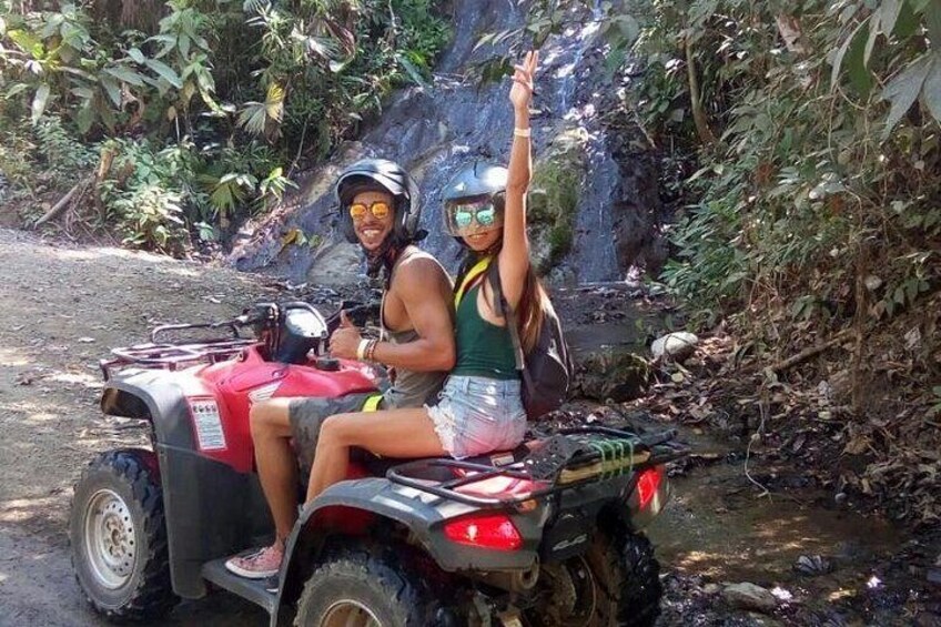 Two people on a red ATV in a forested area with a stream. 2