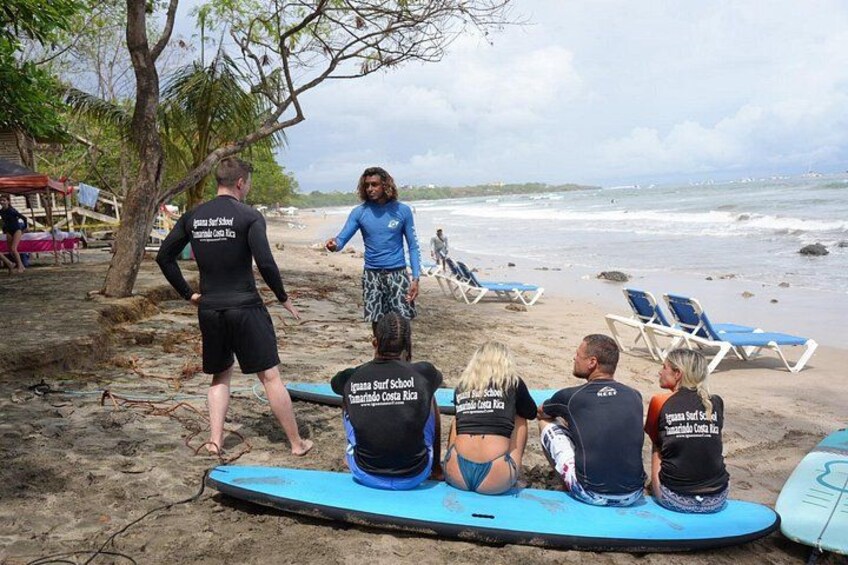 Un groupe de personnes sur une plage, avec des planches de surf, une personne debout qui leur parle, et une tour de surveillance des sauveteurs en arrière-plan. 2