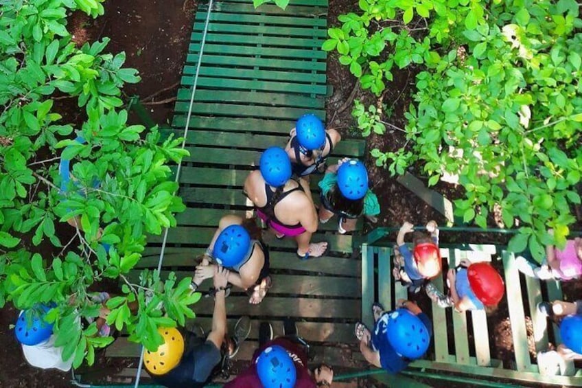 A group of people wearing helmets and harnesses, engaged in a climbing activity on a wooden structure surrounded by greenery. 3