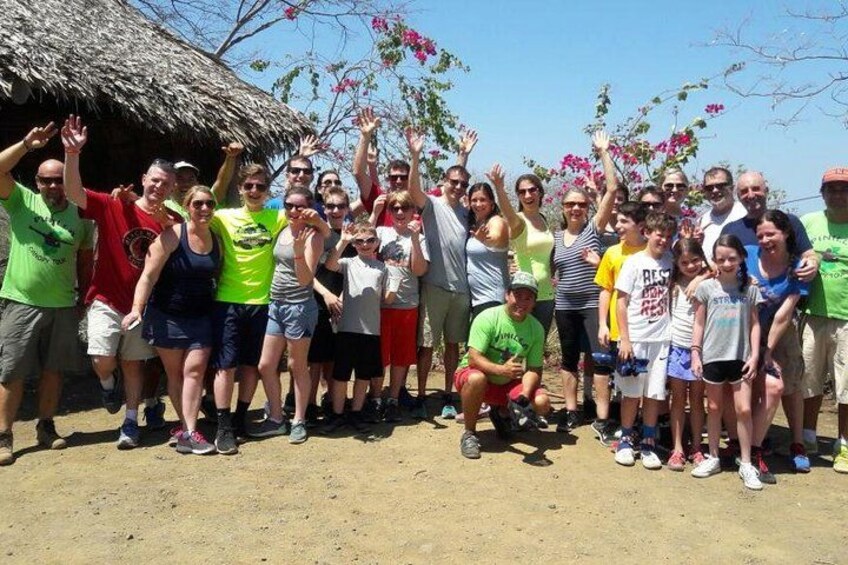 A group of people posing in front of a thatched-roof structure with a clear sky and flowering trees in the background. 1