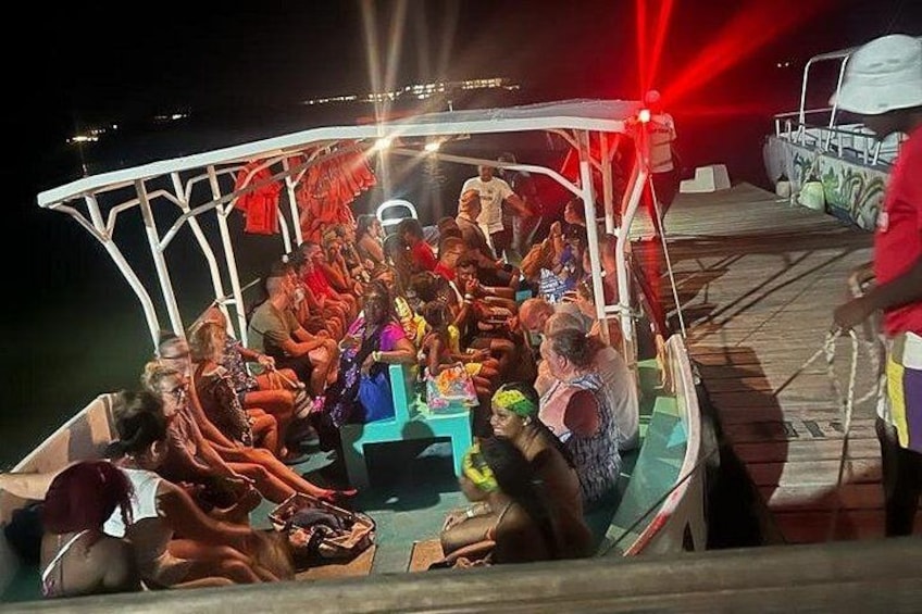A boat with people seated around a table, a person standing on the deck, and a dock in the background. 1