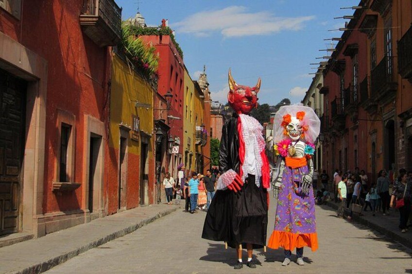 A street with colorful buildings, a person in a red mask, and another in a purple and orange costume. 3