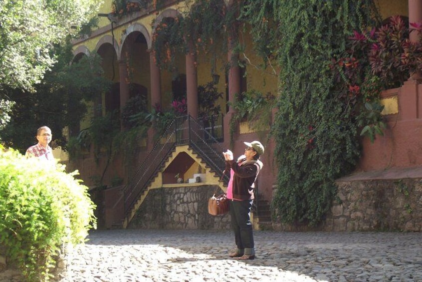 Une personne prend une photo avec un appareil photo dans une cour dallée de pierres, avec un escalier et un bâtiment orné d’arches et de lierre. 3