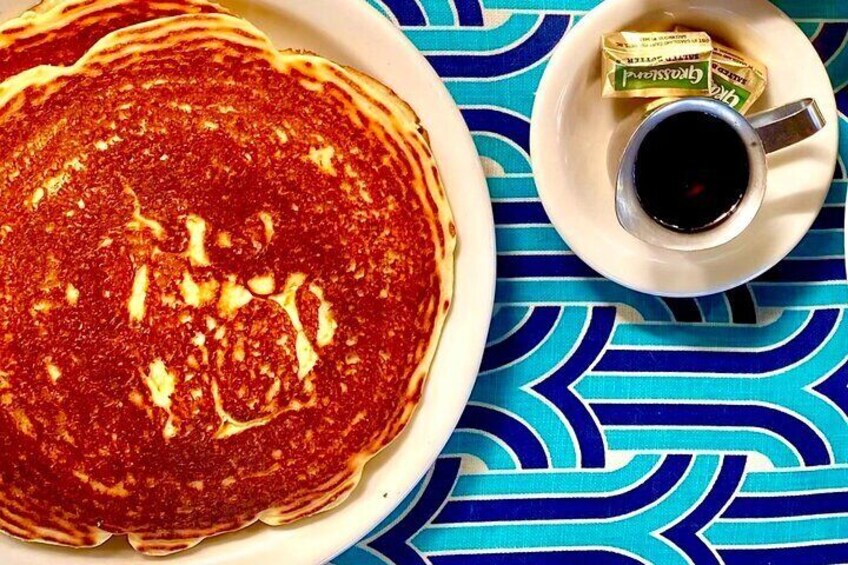A stack of pancakes with syrup, a cup of coffee, and a packet of sugar on a blue patterned tablecloth. 4