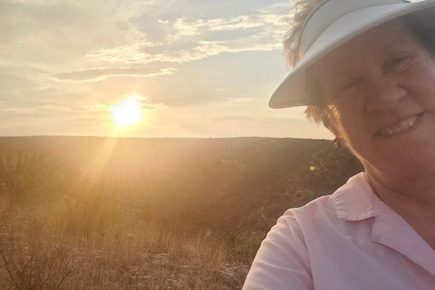 En person med hvid solskærm og pink T-shirt står udendørs med solnedgang i baggrunden. 4