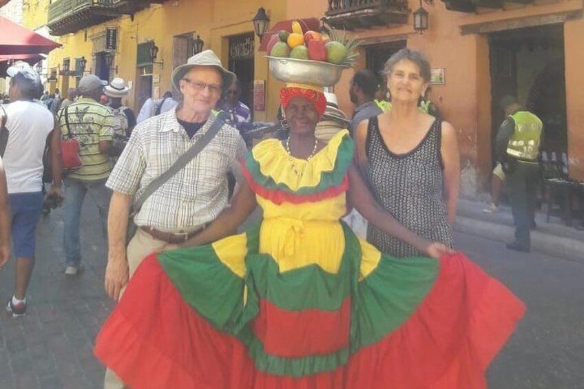 A person in a colorful dress with a headpiece, standing on a street with buildings and people in the background. 4