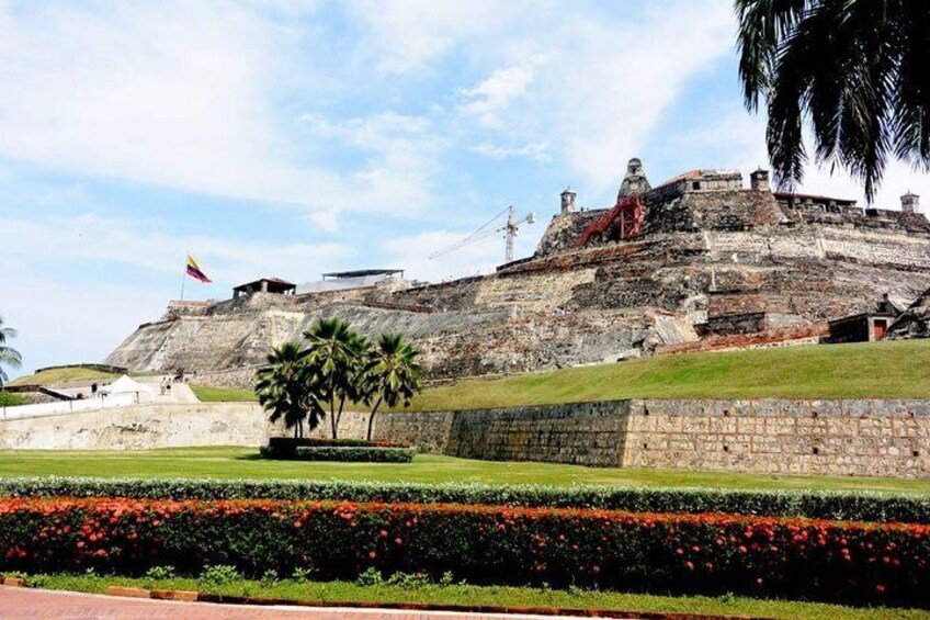 A historical fort with a flag, palm trees, and a flower bed. 1