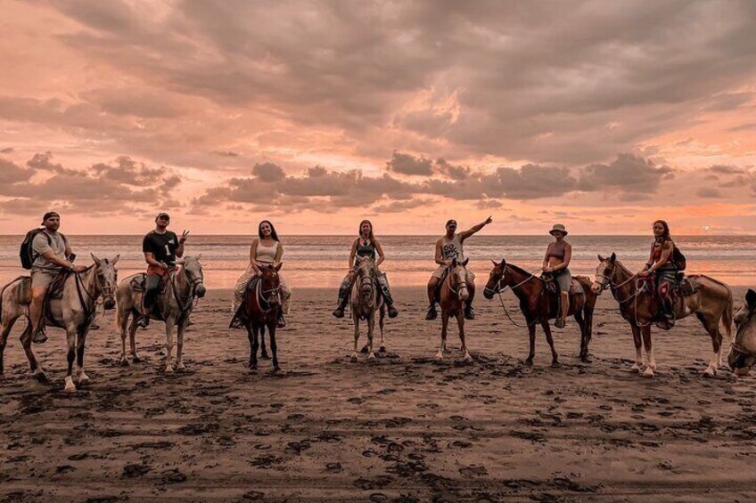 A group of people riding horses on a beach at sunset. 1