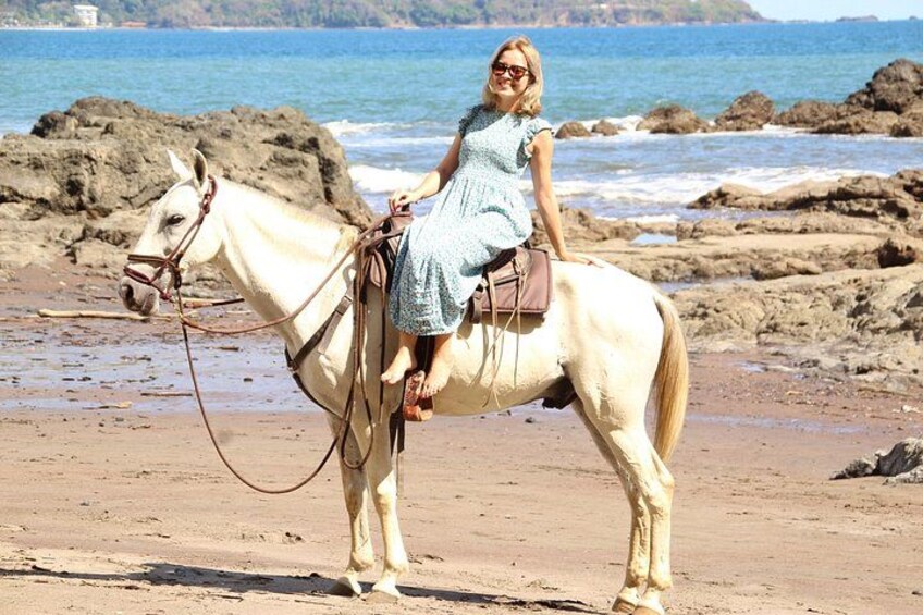 A woman riding a white horse on a sandy beach with rocky outcrops and the ocean in the background. 4
