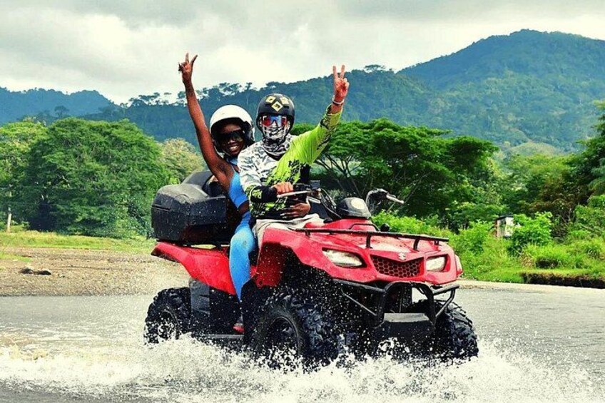 Two people on a red ATV making a peace sign, splashing water, with a mountainous landscape in the background. 2