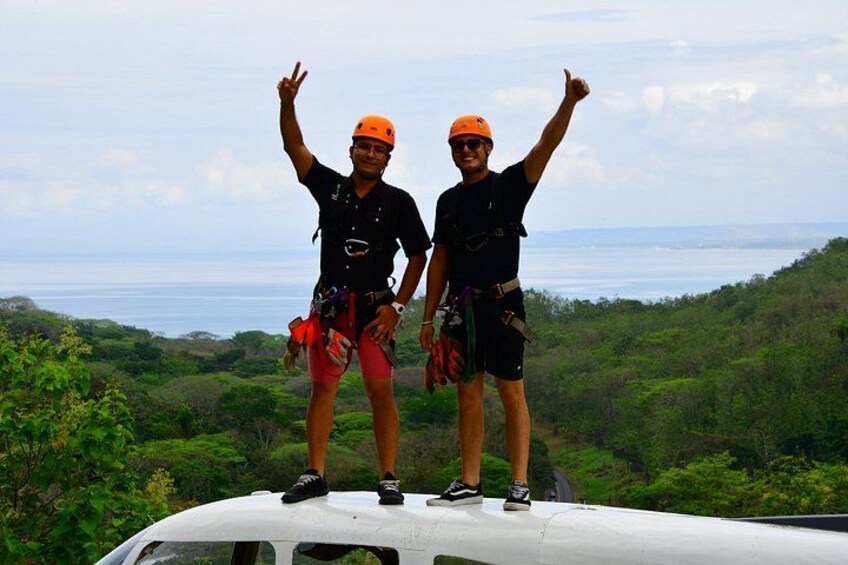 Two people standing on a vehicle with a scenic view of a forest and the ocean in the background. 3