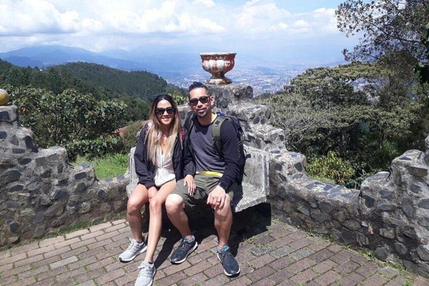 Two people sitting on a stone bench with a view of a valley and a large urn in the background. 1