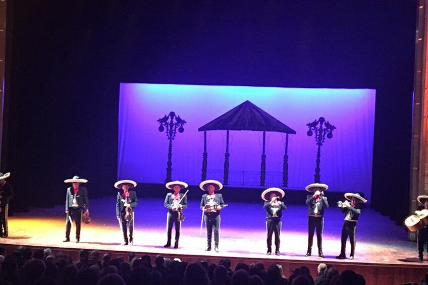 A stage with seven performers in traditional attire, a large screen with a gazebo image, and an audience in the foreground. 5