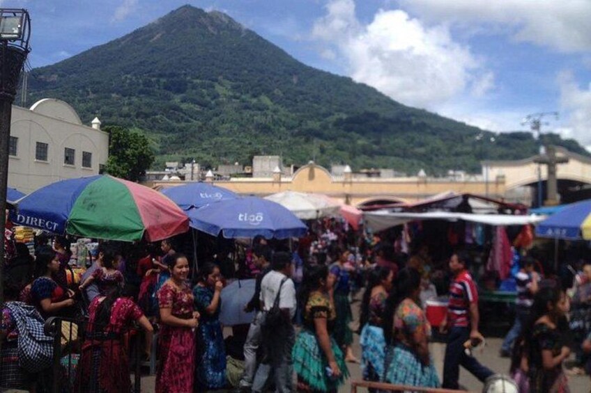 A crowded outdoor market with people and colorful umbrellas, a mountain in the background, and a building with a sign that reads 'DIRECT'. 2