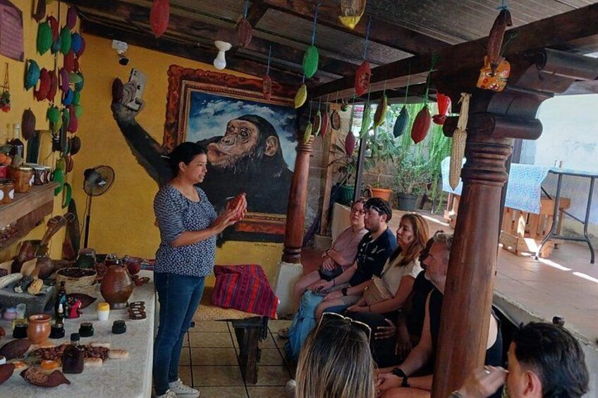 A person standing and gesturing in front of a group of seated individuals in a rustic setting with wooden beams, a mural, and various pottery items. 5