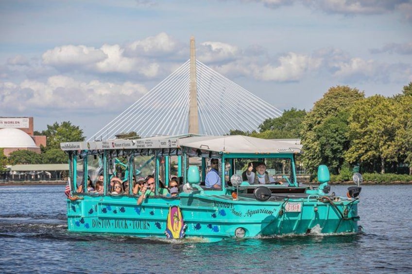 A teal boat with 'Boston Duck Tours' written on it, passengers on board, and a bridge in the background. 1