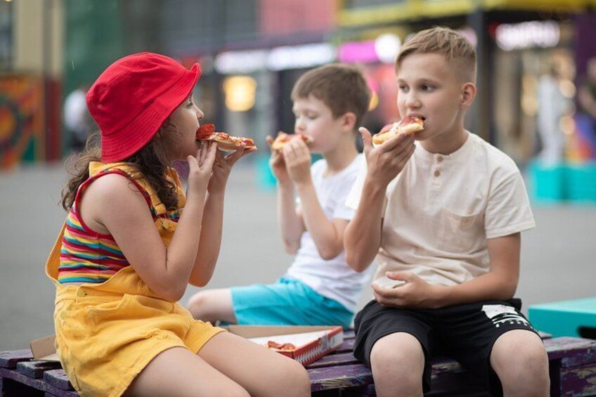 Three children sitting on a bench eating pizza in an urban setting. 1