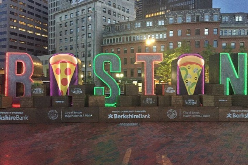 Illuminated letters spelling 'BOSTON' with pizza designs on each letter, set against a cityscape backdrop. 3